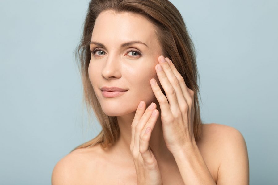 Beautiful millennial Caucasian naked woman isolated on blue studio background touch soft healthy face skin pleased with beauty procedures, pretty blonde female satisfied with clinic facial injection