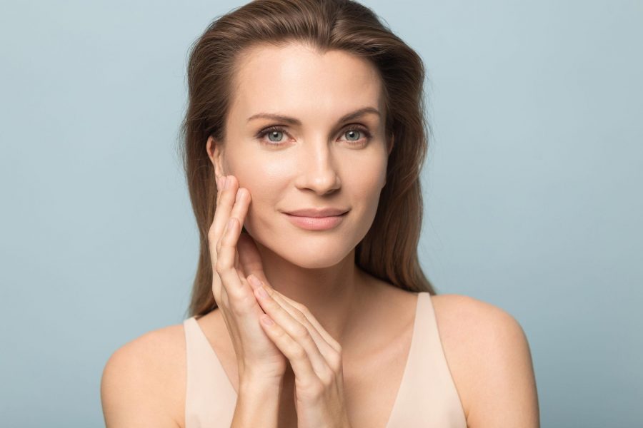 Close up portrait of beautiful young woman isolated on blue studio background touch clean healthy skin, pretty millennial female model look at camera satisfied with moisturized hydrated face treatment